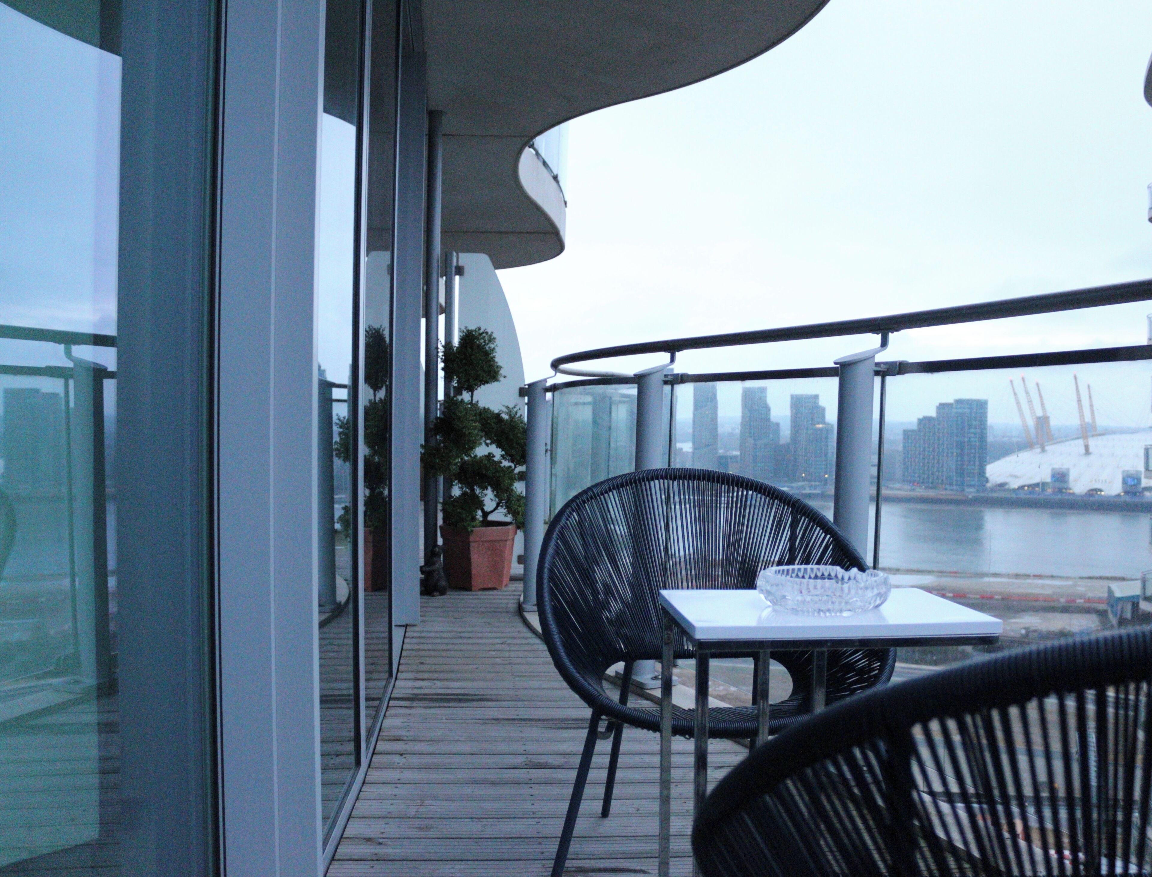 A balcony with a table and chairs, a potted plant, and a view of a city skyline and river.