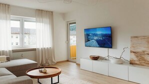 Ein modernes Wohnzimmer mit einer Couch, einem runden Couchtisch, einem an der Wand montierten Fernseher und Blick auf die Stadt durch das Fenster.