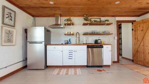 A kitchen with white cabinets, a stainless steel dishwasher, and wooden shelves above the counter. - Quaint Cottage Downtown Buckner (Buckner)