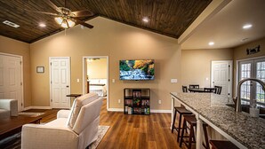 A spacious living room with a high ceiling, wooden flooring, a comfortable beige armchair, a television mounted on the wall, and a kitchen area with a granite countertop and bar stools. - The Red Door Cottage (Dahlonega)