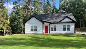 A single-story house with a red door, white siding, and a black roof, surrounded by a well-maintained lawn and trees. - The Red Door Cottage (Dahlonega)