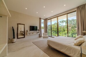 A modern bedroom with a large bed, a TV on a built-in shelf, a potted plant, and a view of trees through large windows. - Modern 4-bedroom  4- bathroom villa with private pool in Tulum.  (Tulum)
