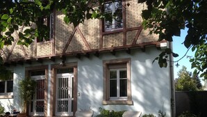 Uma casa de dois andares com fachada branca, chaminé de tijolos e uma pequena varanda frontal com duas cadeiras.