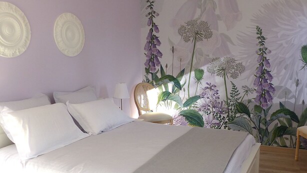 Quarto com cama, almofadas, mesa de cabeceira, abajur e papel de parede floral.