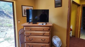 Une chambre dotée d’une commode en bois, d’un téléviseur à écran plat, d’un ventilateur et d’une porte donnant sur un balcon avec vue sur un paysage montagneux.