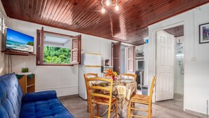 Una sala acogedora con un sofá azul, una mesa de comedor con sillas, un televisor montado en la pared y un ventanal con vistas a la vegetación.