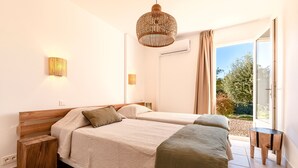 A modern bedroom with two beds, a wooden side table, a small rug, and a large window with a view of greenery. - Casa Vinti - Vue mer et montagne, piscine (Grosseto-Prugna)