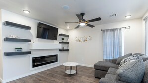Ein modernes Wohnzimmer mit einem grauen Sofa, einem weißen runden Couchtisch und einem flachbildfernseher an einer weißen Wand. Es gibt einen Deckenventilator, Einbauleuchten und ein Fenster mit Vorhängen.
