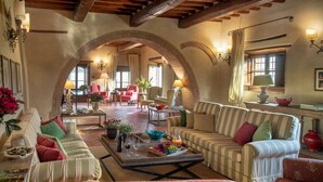 A spacious living area with a high ceiling, exposed wooden beams, and a large arched doorway leading to another room. There are multiple seating options including striped sofas and armchairs, a coffee table with a fruit platter, and a fireplace.