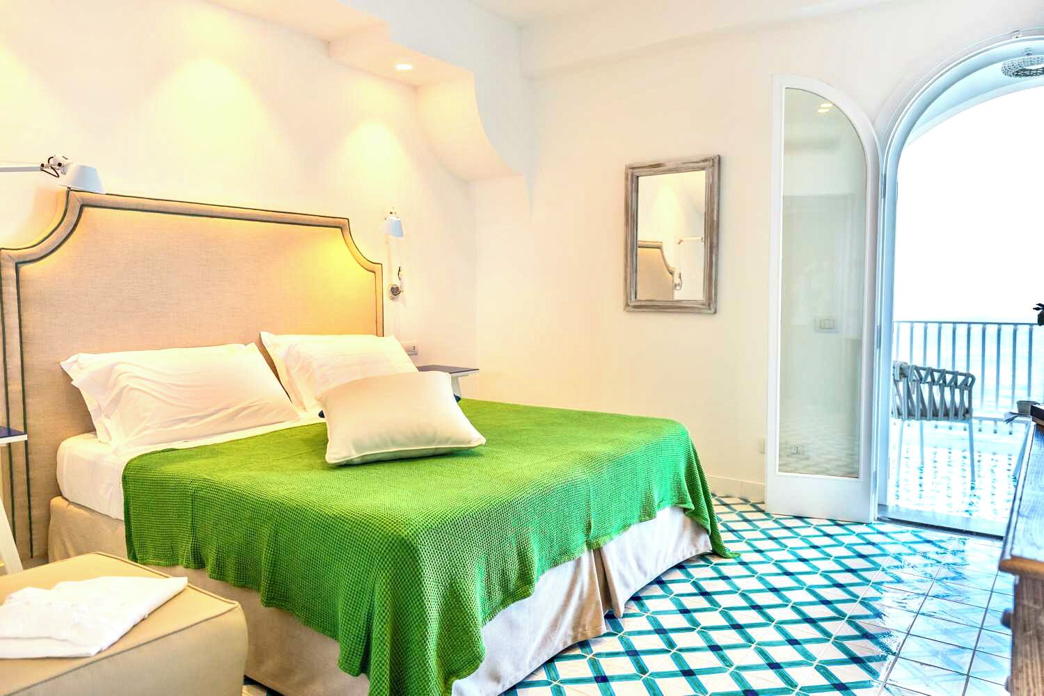 A hotel room with a bed, a nightstand, a chair, a balcony with a railing, and a checkered floor.