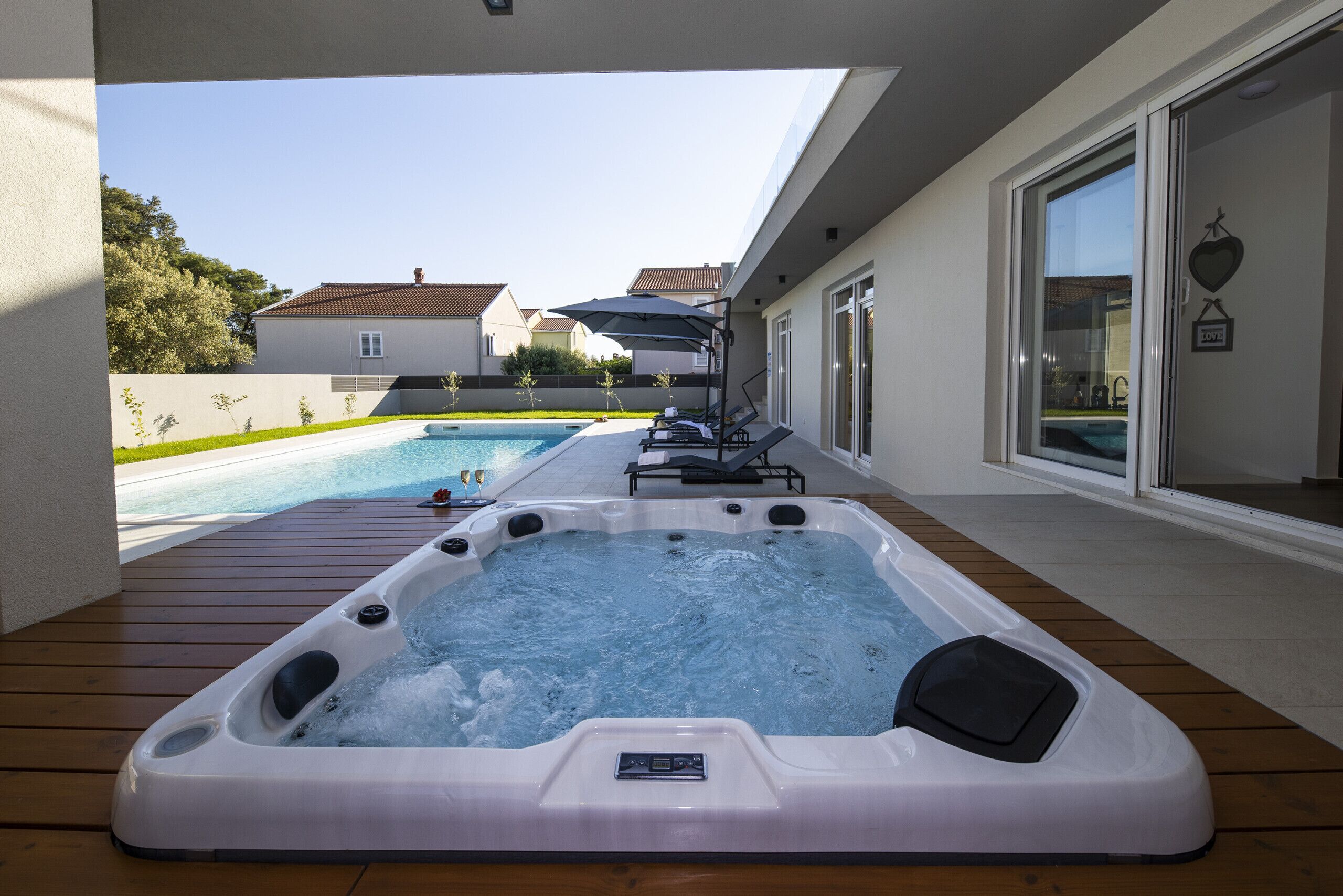 A hot tub on a wooden deck with a pool in the background.