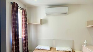 A single bed with a patterned bedspread, two pillows, a bedside table with a lamp, and an air conditioning unit mounted on the wall.