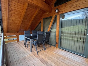 Eine Holzterrasse mit Tisch und Stühlen, große Schiebeglasfenster und Blick auf einen umzäunten Garten.