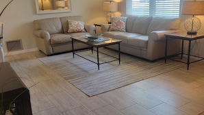 A living room with a beige sofa, two coffee tables, and a large window with blinds. - Charming 3-bedroom house in quaint Venice with heated pool! (Venice)
