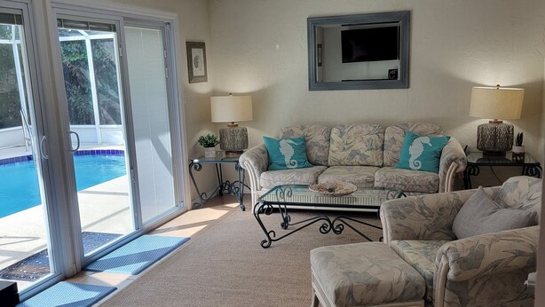 A living room with a pool view, a sofa with teal cushions, a TV mounted on the wall, and a patterned rug. - Charming 3-bedroom house in quaint Venice with heated pool! (Venice)