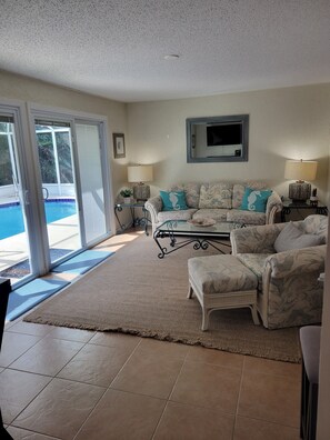 Ein Wohnzimmer mit Blick auf einen Pool, eine Couch mit türkisfarbenen Kissen, ein an der Wand montierter Fernseher und ein gemustertes Teppich.