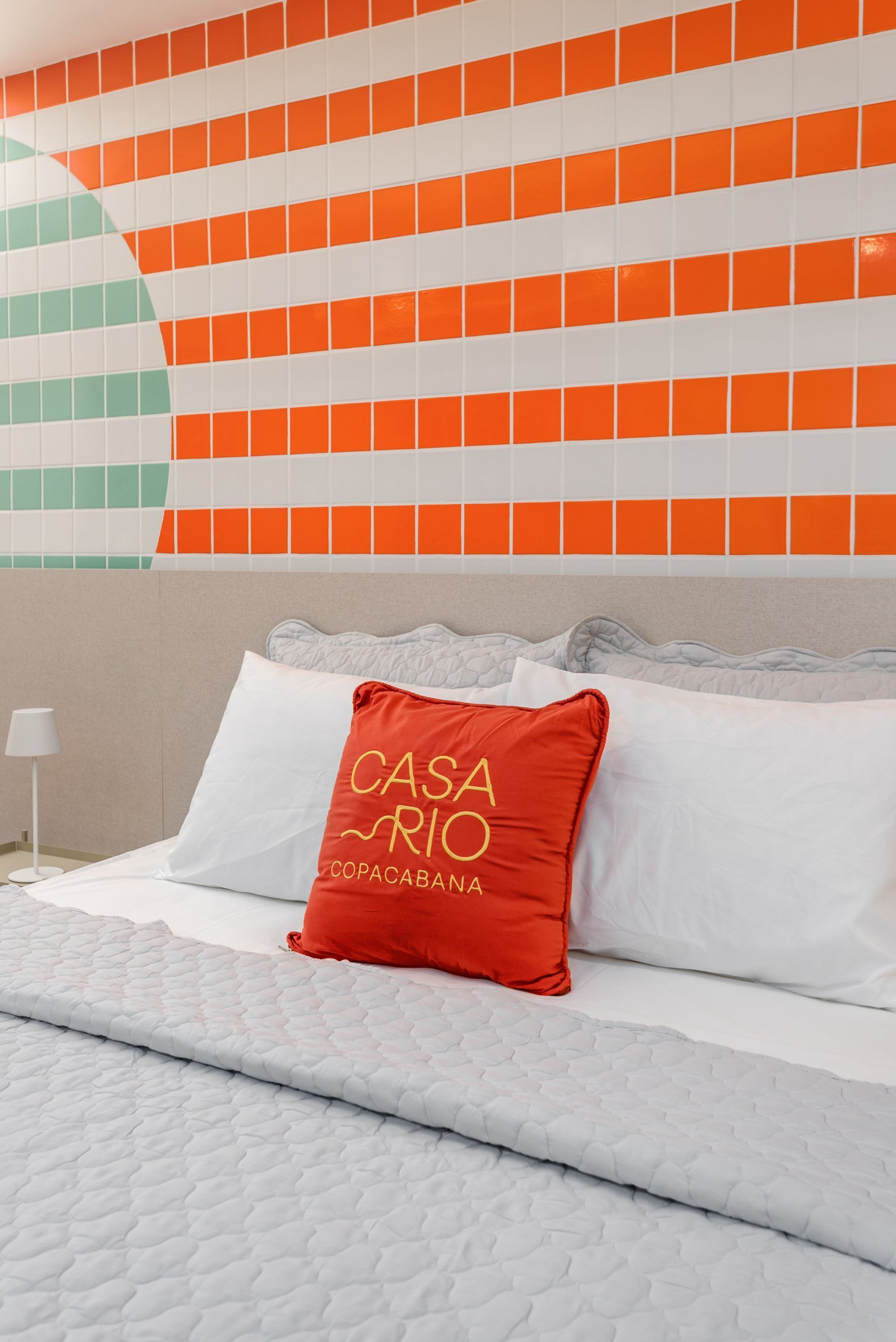 Une chambre d’hôtel avec un lit, un oreiller rouge orné d’un motif, et un mur recouvert de carreaux orange et blancs.