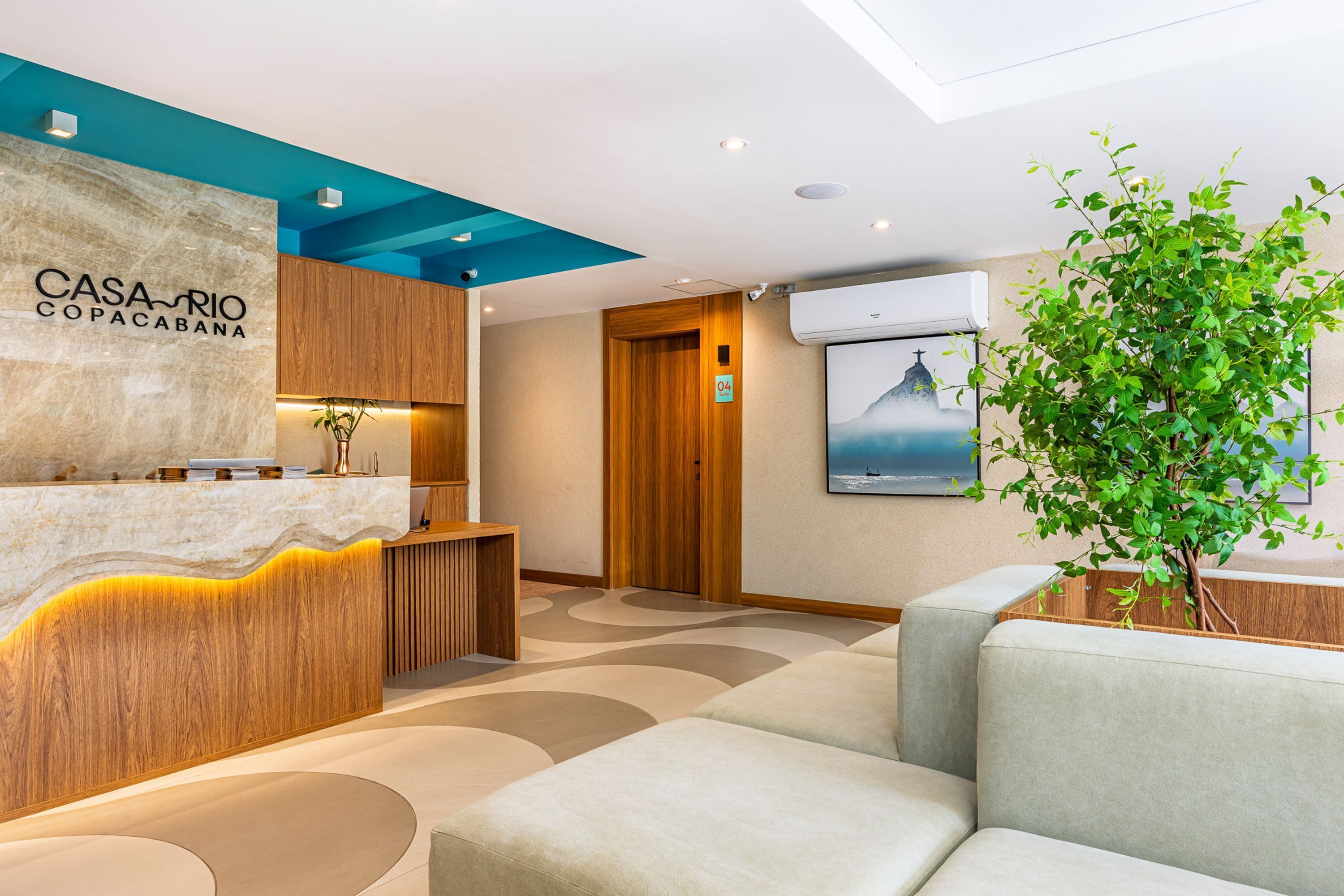Un espace d’accueil moderne, avec un comptoir de réception en bois, une grande plante en pot et un coin salon avec des canapés.