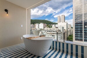 A modern bathroom with a freestanding bathtub, a tiled floor, and a large window offering a city view.