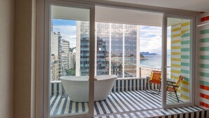 A modern bathroom with a large bathtub, a window with a view of a beach and buildings, and a striped floor.