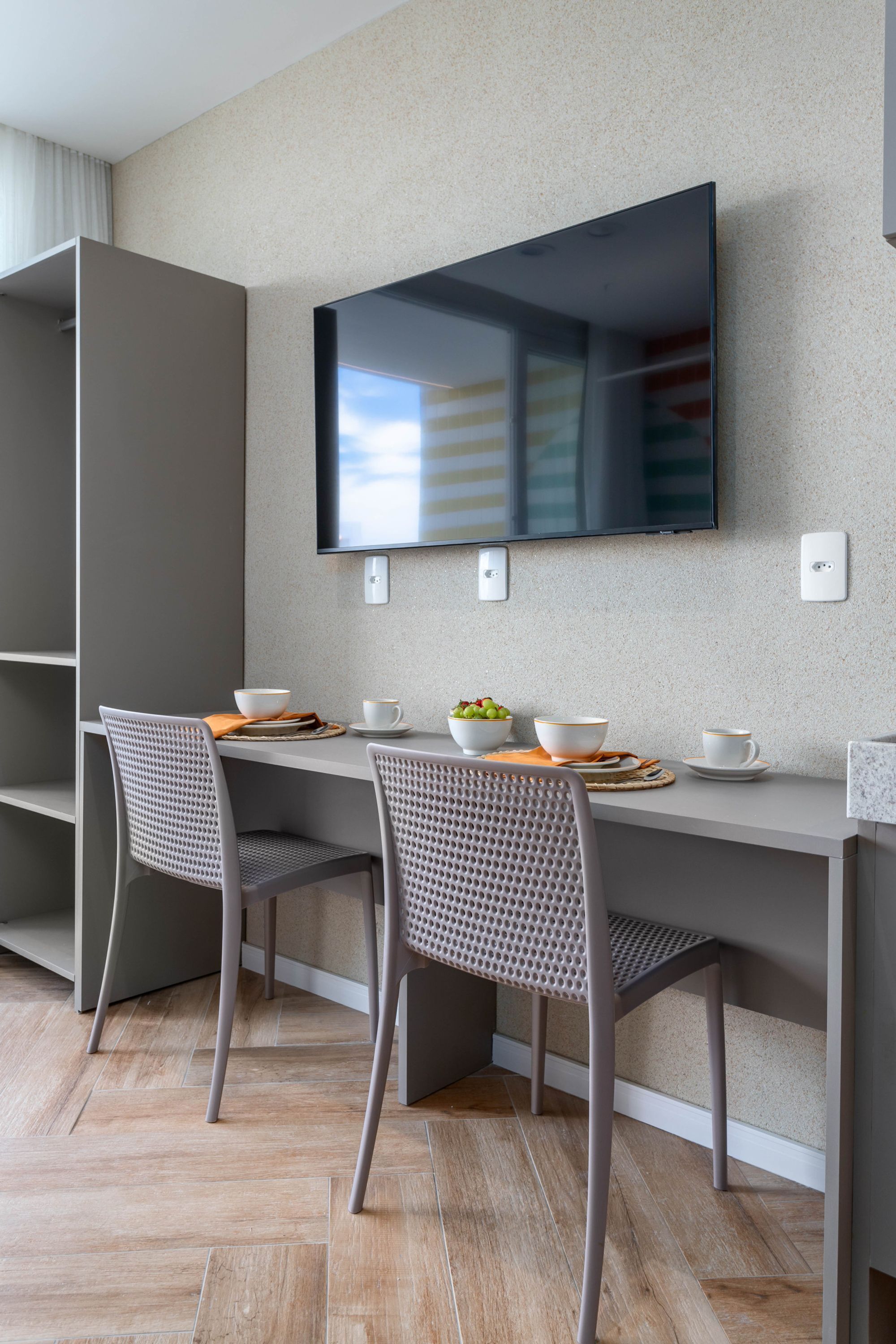 Ein moderner Essbereich mit einem an der Wand befestigten Flachbildfernseher, zwei Stühlen, einem Tisch mit Schüsseln und Tassen sowie Holzboden.