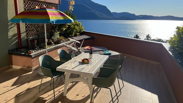 Een terras met een tafel, stoelen en een kleurrijke parasol, met uitzicht op een meer met bergen op de achtergrond.