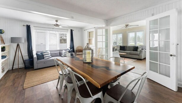 A spacious living area with a wooden dining table, white chairs, a sofa, and a large window. - Sleeps 8-Cozy 3-Bedroom Cottage, 2 Blocks from the Strip, 10 Minutes from Spire (Geneva)