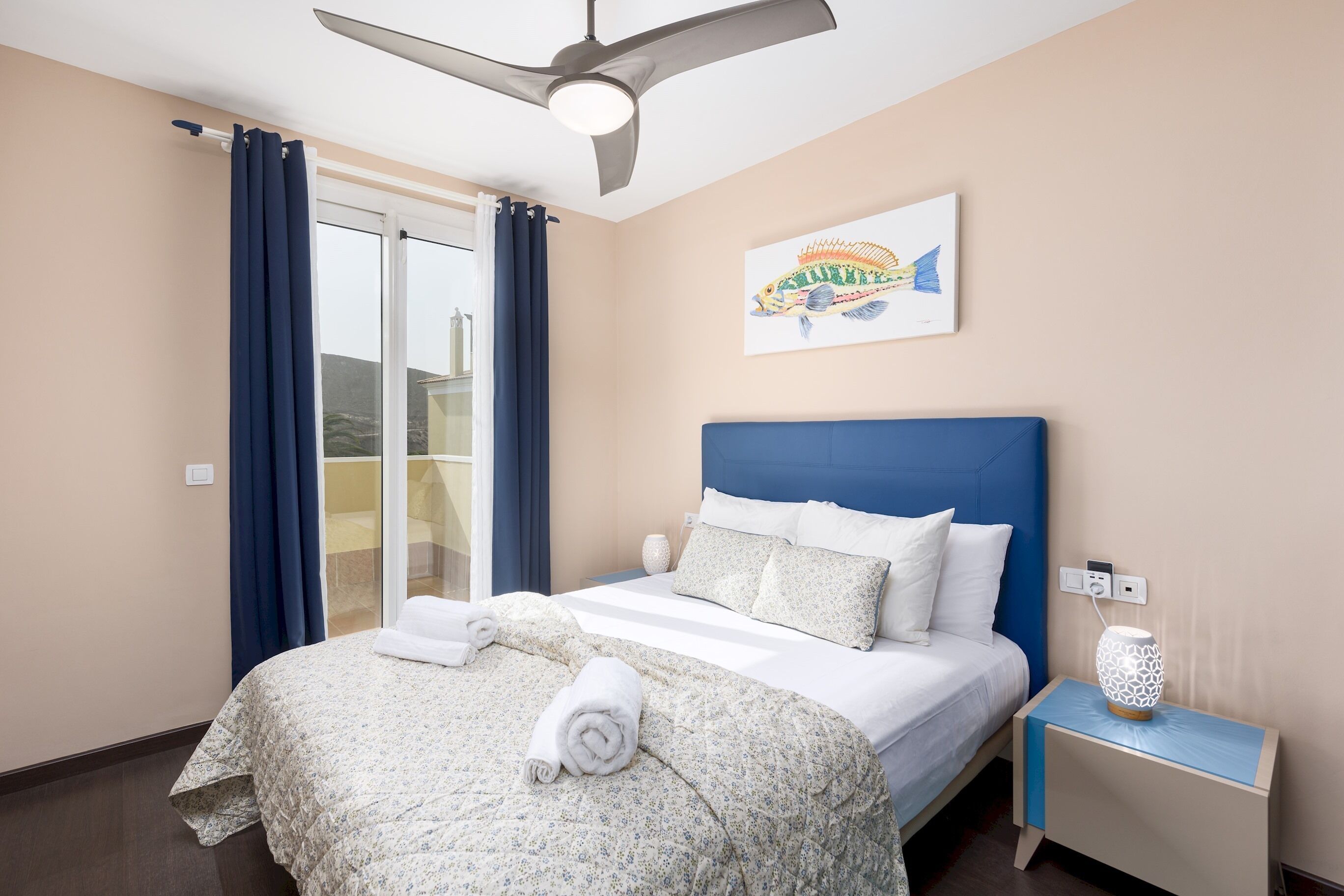 Een hotelkamer met een bed, nachtkastje, plafondventilator, raam met gordijnen en een kunstwerk aan de muur.
