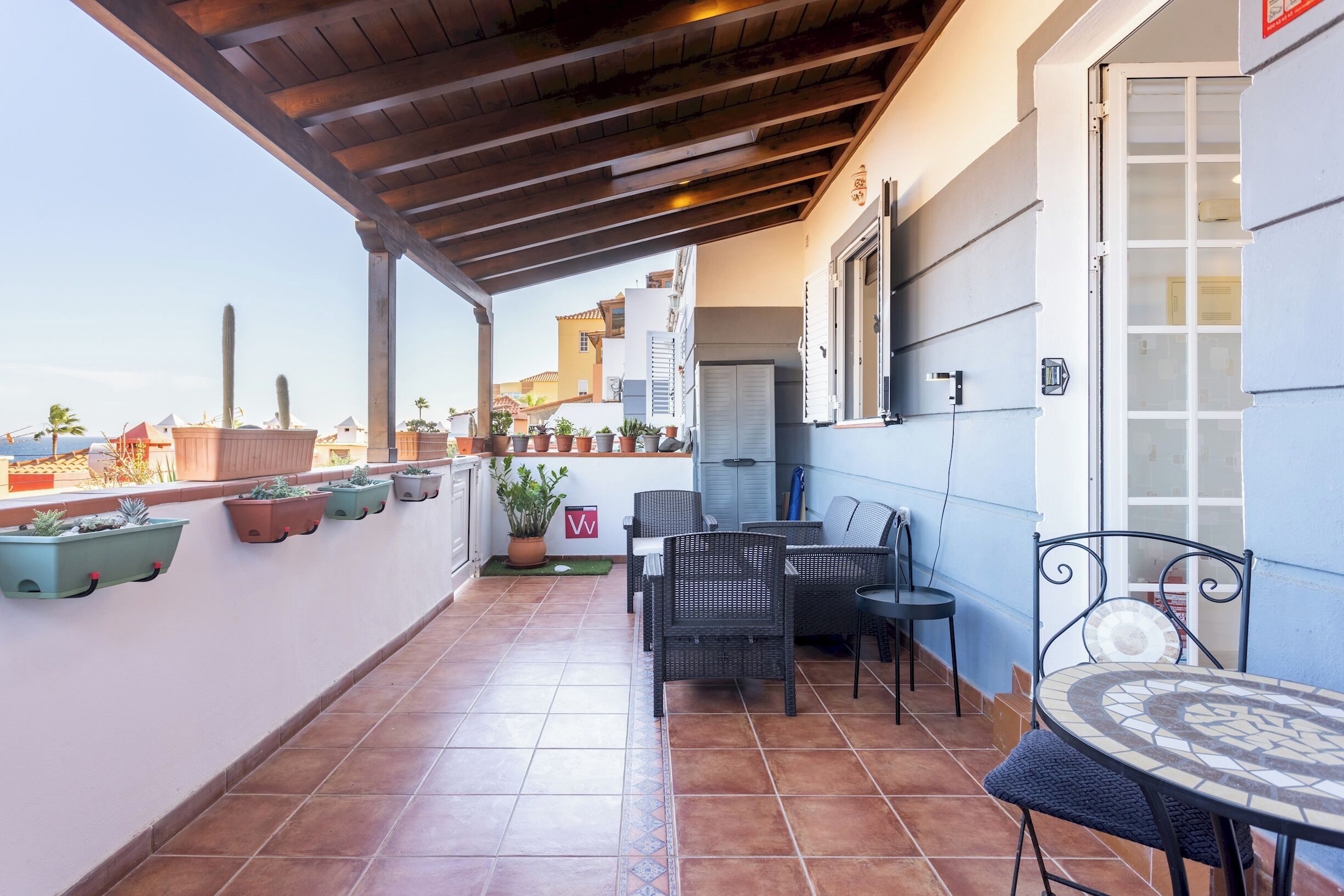 A covered patio with outdoor furniture, potted plants, and a view of the city.