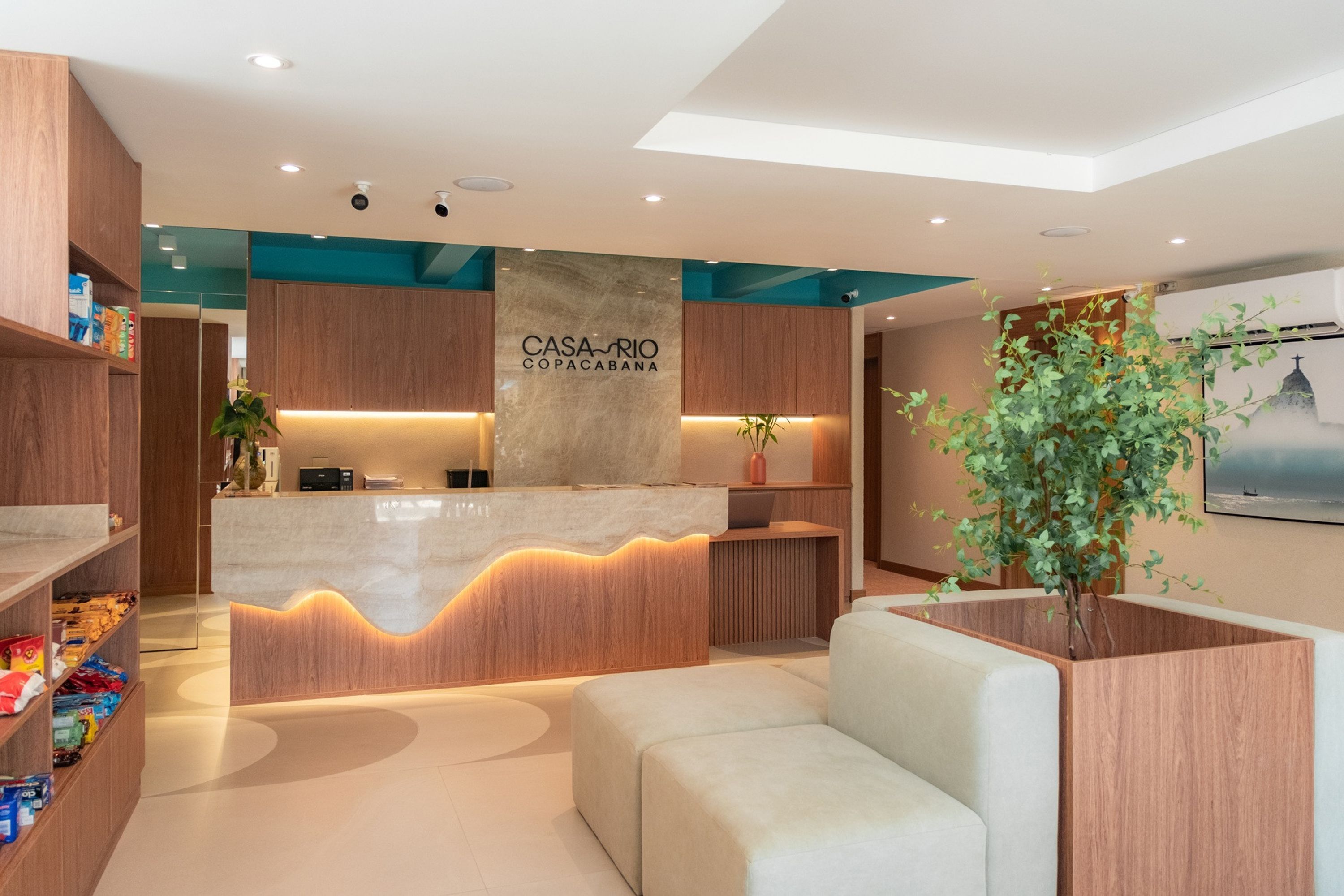 A modern reception area with a wooden reception desk, a large sign reading "CASA-RIO COPACABANA," and a seating area with a potted plant.
