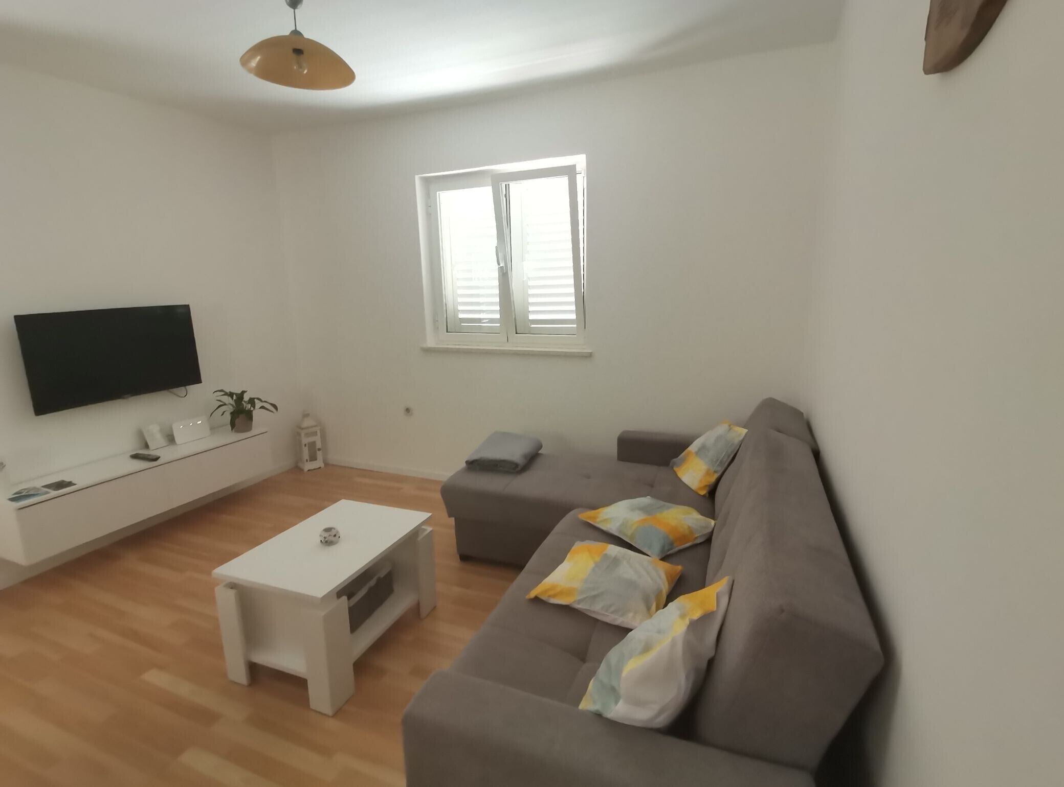 A modern living room with a flat-screen TV, a white console table, a grey sofa with patterned cushions, a small white table, and a window with blinds.