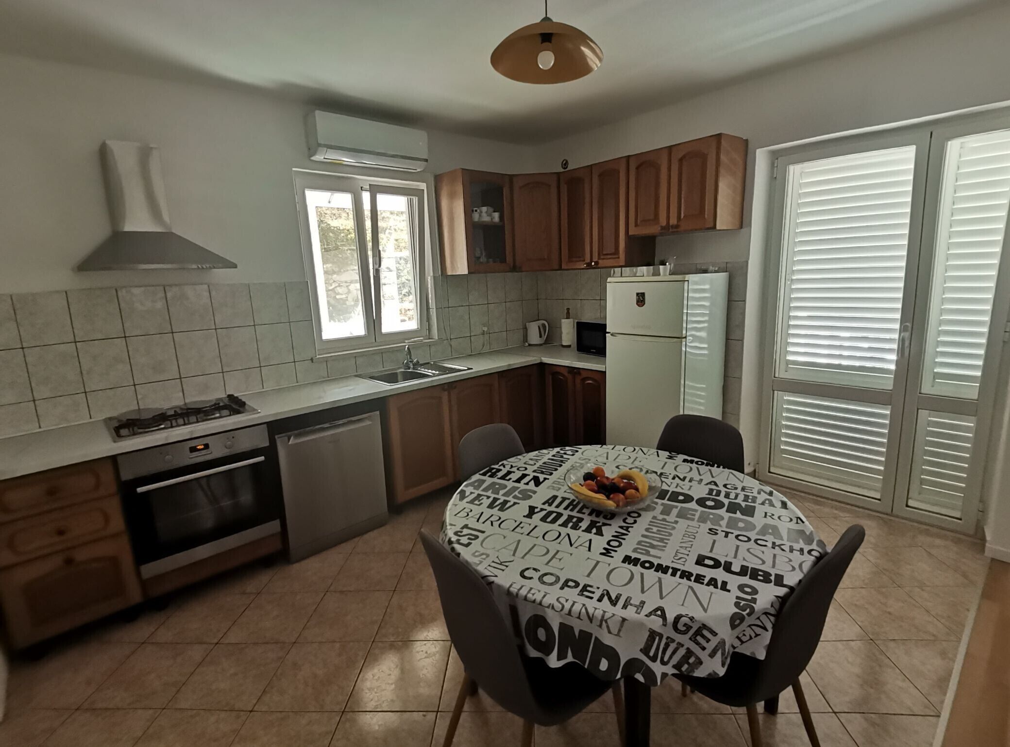 Een moderne keuken met houten kastjes, een gasfornuis en een ingebouwde oven. Er staat een eettafel met een rond tafelkleed waarop stadsnamen staan.