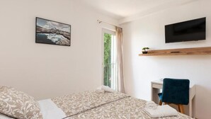 A bedroom with a bed, a desk, a chair, a TV, and a window with a view of greenery.