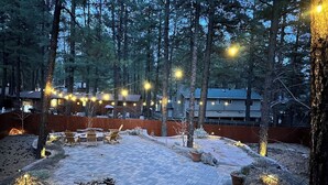 Un patio al aire libre con suelo empedrado, mobiliario y luces colgantes, rodeado de árboles.
