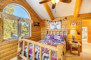 A log cabin bedroom with a wooden bed, a window with a view of trees, and a ceiling fan. - Private Fishing Pond: Mtn-View Home in Florissant! (Florissant)