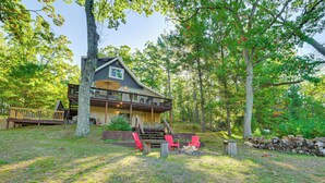 A cabin with a deck, surrounded by trees and a fire pit.
