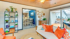 Een ruime woonkamer met houten plafond, een plafondventilator, een bank met oranje kussens, een boekenkast en uitzicht naar buiten.