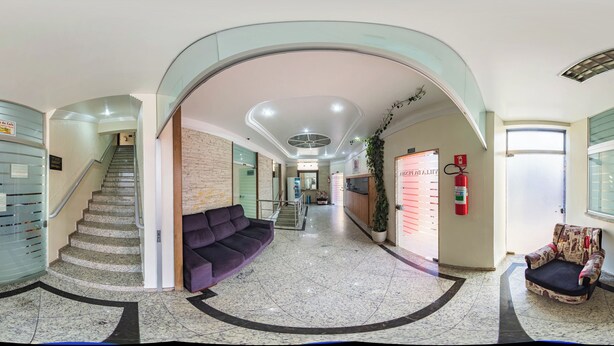 Un vestíbulo de hotel con un muro de cristal curvo, una escalera, un sofá morado y un extintor.