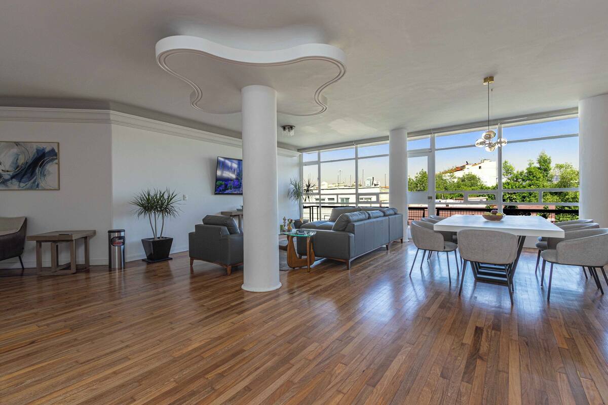 A spacious living area with a wooden floor, a gray sofa, a dining table with chairs, and large windows offering a view of the city.