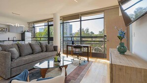 Ein modernes Wohnzimmer mit einer Couch, einem Couchtisch und einem Essbereich mit Tisch und Stühlen. Große Fenster bieten Blick auf die Stadtlandschaft.