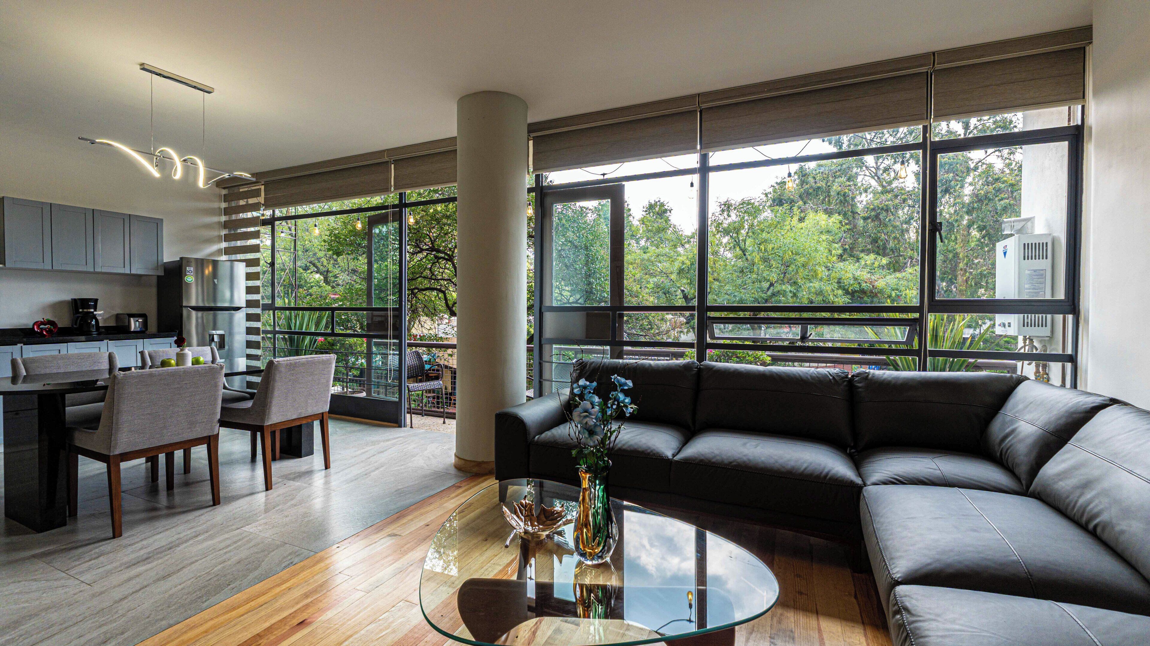 Un espacio habitable moderno con una mesa de centro de vidrio, un sofá de cuero negro y vistas a zonas verdes a través de amplios ventanales.