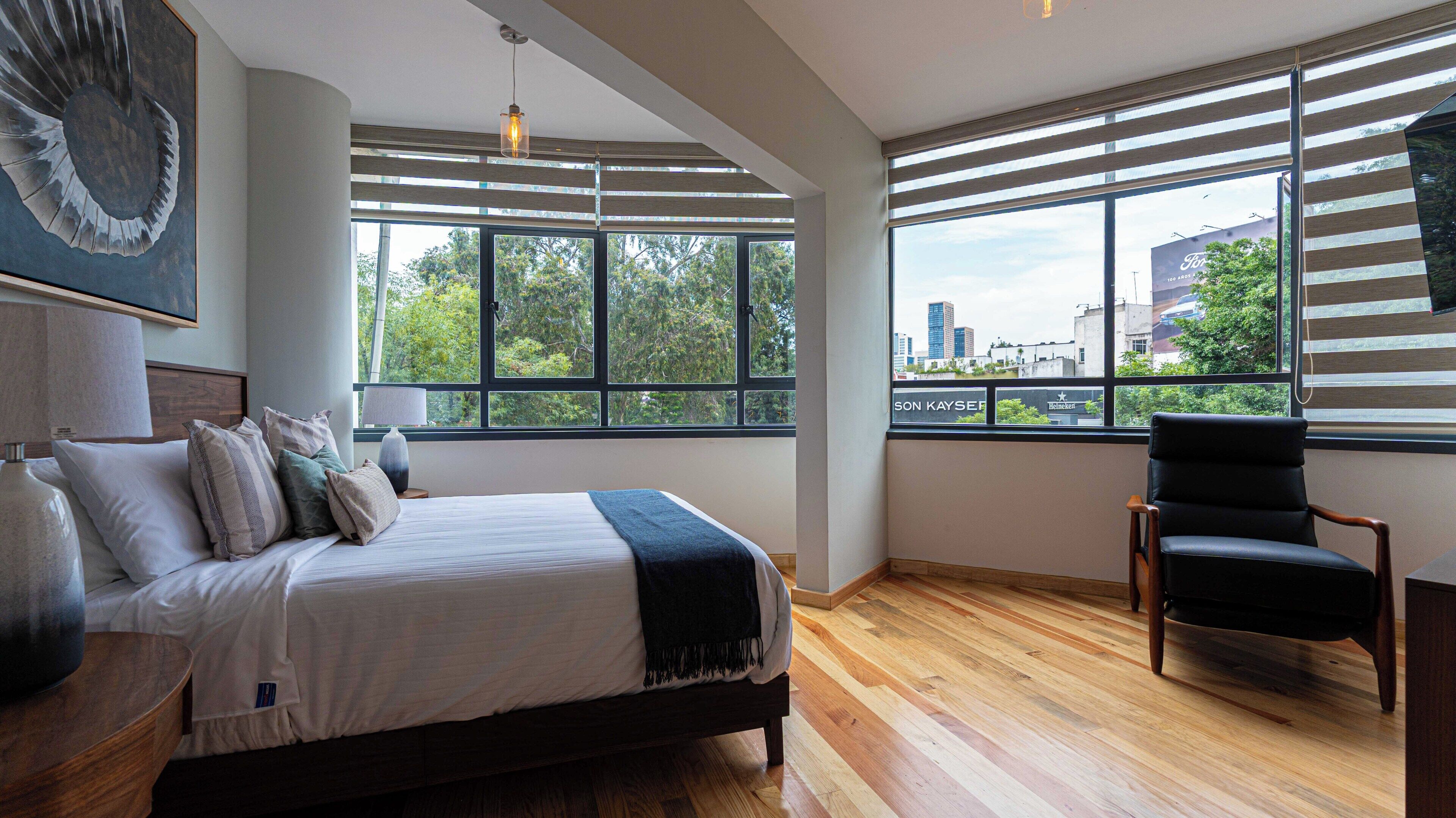 Un dormitorio moderno con una cama grande, una silla y vistas de la ciudad a través de amplios ventanales.