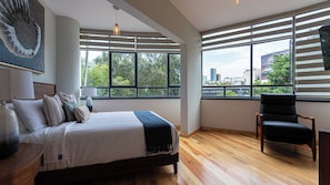 Un dormitorio moderno con una cama grande, una silla y vistas de la ciudad a través de amplios ventanales.