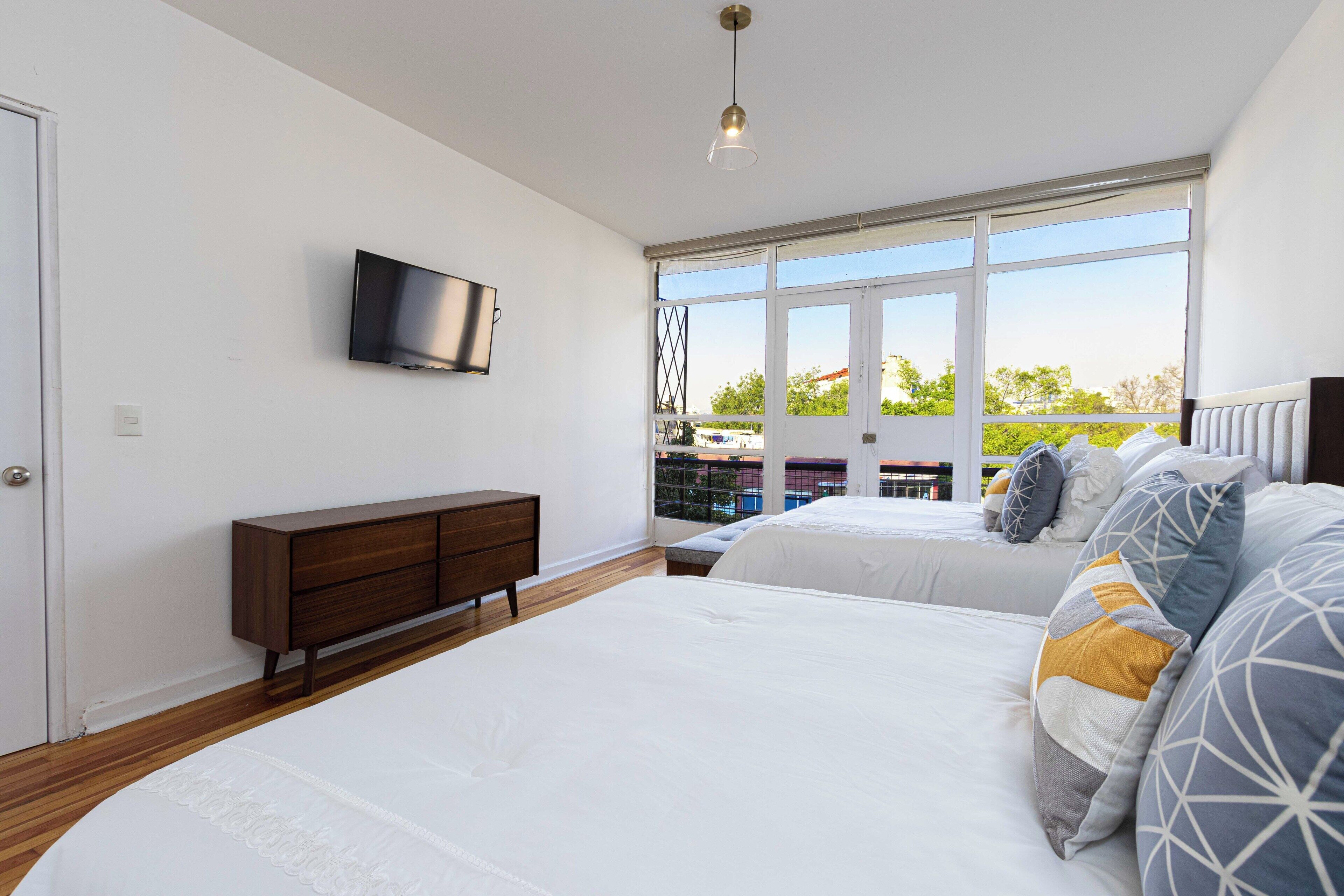 A bedroom with two beds, a TV mounted on the wall, and a large window providing a view of the outdoors.