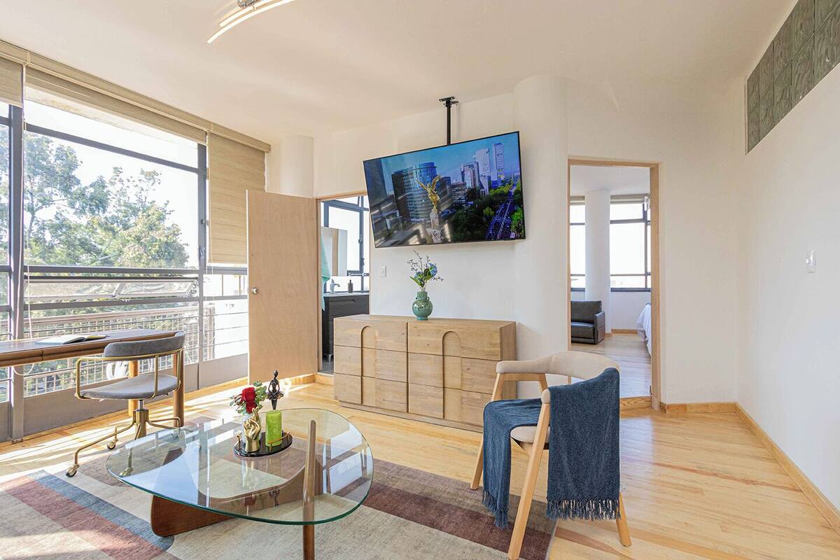 A spacious living room with a glass coffee table, a sofa, a TV mounted on the wall, and a view of the city through large windows.