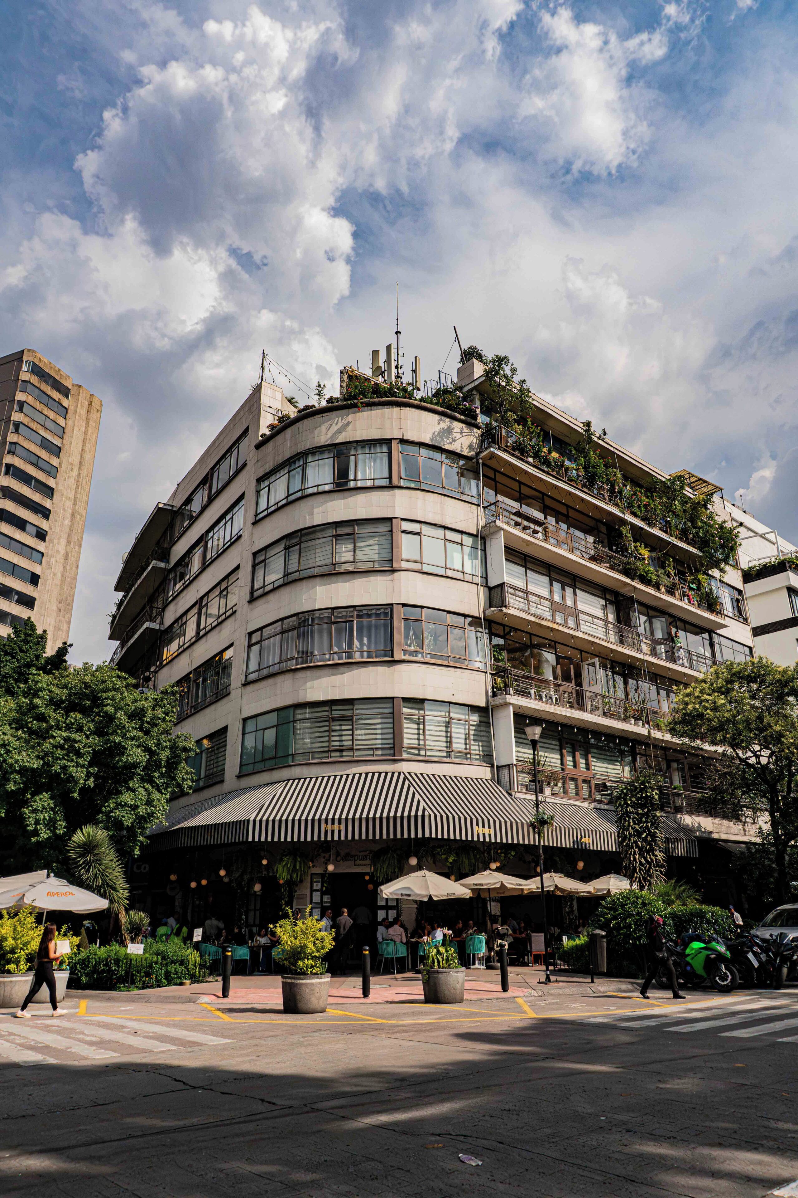 Un hotel de varios pisos con fachada curva, área de estar al aire libre con sombrillas y vista a la calle.