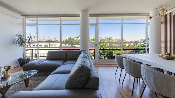 Ein modernes Wohnzimmer mit einem Ecksofa, einem Glastisch und einem Essbereich mit weißem Tisch und Stühlen.
