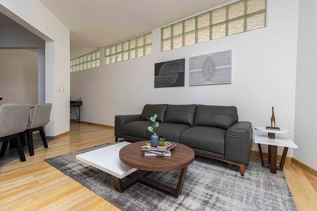 A modern living room with a grey sofa, a round coffee table, and a small white side table. There are two abstract paintings on the wall and a window with patterned blinds.