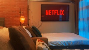 A hotel room with a brick wall, a bed with multiple pillows, a wall-mounted TV displaying the Netflix logo, and a hanging light fixture.