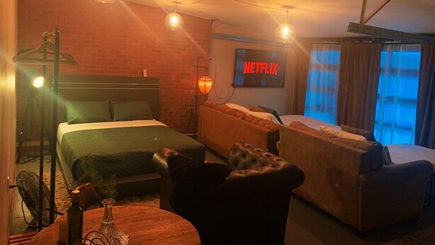A hotel room with a bed, a sofa, a table with a bottle, and a TV displaying "NETFLIX".